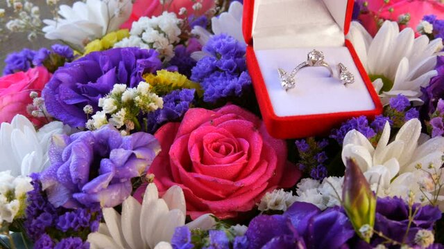 Gift present red box with a set of engagement ring and earrings on the background of a multi-colored beautiful bouquet of flowers. Marriage proposal. White gold or silver with diamond stone
