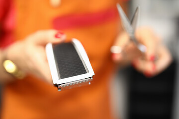 Close-up of woman holding scissors and shaving machine. Professional hairdresser working in beauty salon. Macro shot of female with red fresh manicure. Barber shop concept