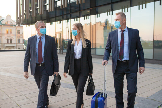 Business Men And Woman In Face Masks Travelling With Luggage, Walking Outdoors With Attendant, Talking To Each Other. Front View. Business Trip And Epidemic Concept