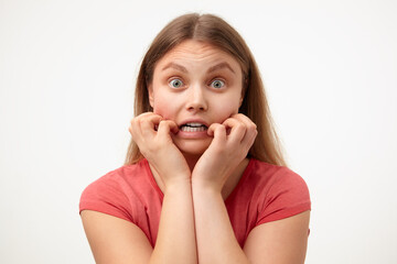 Frightened young green-eyed blonde woman with loose long hair holding her face while looking scaredly at camera, standing over white background in casual clothes