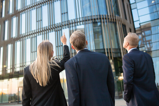 Business Professionals In Suits Looking At Office Building Facade, Pointing Up, Discussing Real Property. Back View, Low Angle. Commercial Real Estate Concept