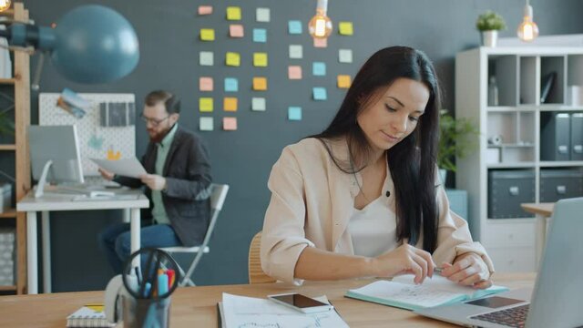 Businesswoman young brunette is using laptop typing then taking notes in notebook, male coworker is working in background. Job and people concept.