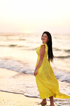 A girl in a long dress with long black hair walks at dawn along a deserted beach along the line of the surf. 