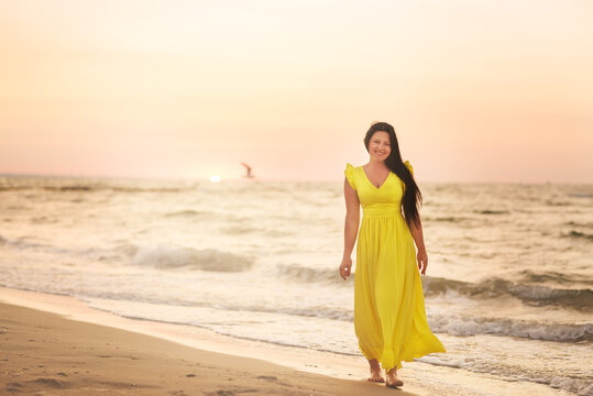 A girl in a long dress with long black hair walks at dawn along a deserted beach along the line of the surf. 