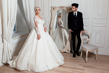the bride and groom in a light studio are standing near the antique mirror and window.