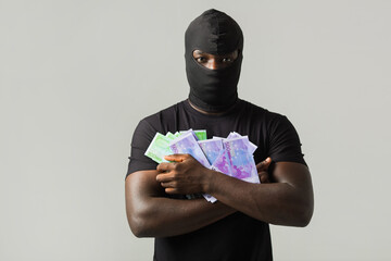 young african man in a black robber mask, and in a black t-shirt on a gray background with euro in hands