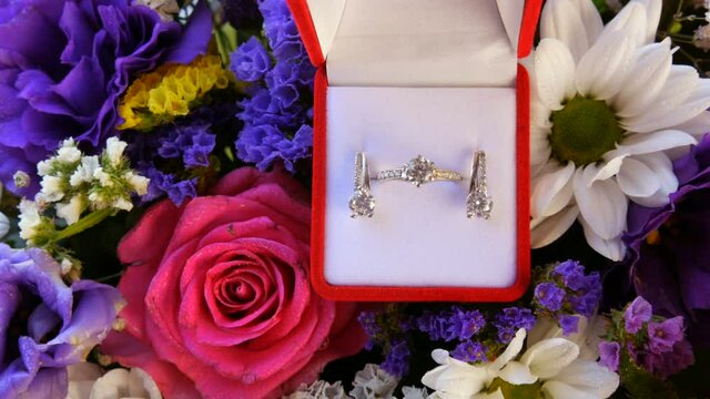 Gift present red box with a set of engagement ring and earrings on the background of a multi-colored beautiful bouquet of flowers. Marriage proposal. White gold or silver with diamond stone