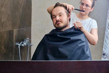 A woman cuts a man's hair at home in the bathroom, reflection in the mirror, copy space