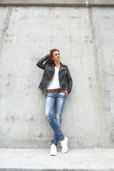 Obraz premium Young cute woman is standing by the wall in a leather jacket, jeans and sneakers.