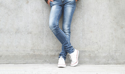 Shapely female legs in sneakers and jeans near a concrete wall.