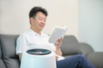 An Asian middle-aged man reading in the living room with an air purifier on. Blur background.