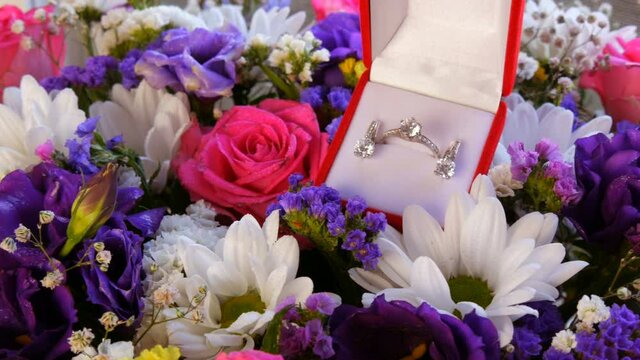 Gift present red box with a set of engagement ring and earrings on the background of a multi-colored beautiful bouquet of flowers. Marriage proposal. White gold or silver with diamond stone