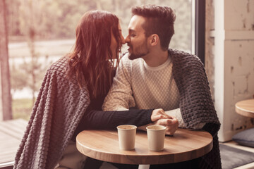 Focus on cups. Close up portrait of lovely couple sitting in the cafe and flitring, kissing, hugging. Attractive husband and wife in coffee shop have a lunch after distance work.