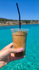 Woman's hand holding Coffee Frappe (Iced Coffee) in clear plastic cup with straw by the sea