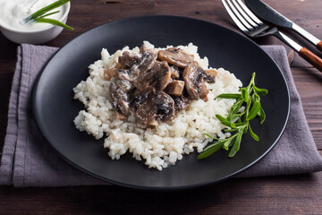 Risotto with mushrooms on a black plate. Boiled white rice with champignons in cream sauce. Dark wooden background.