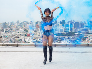Young sexy woman dncer in a blue colored smoke outdoors at the top of a high-rise. Female dancer using a firework as a prop in her dance.