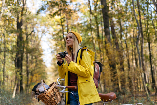 Beautiful smiling woman wearing yellow raincoat and hipster hat riding bicycle in autumn forest. Inspiration, healthy lifestyle, travel concept. Photographer holding camera, taking pictures in park