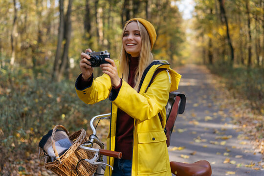 Photographer holding retro camera, taking pictures in park. Beautiful smiling woman wearing yellow raincoat and hipster hat walking in autumn forest. Inspiration, healthy lifestyle, travel concept