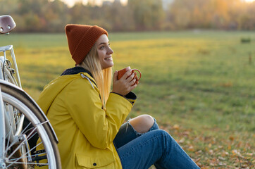 Beautiful smiling woman sitting on grass in park, holding cup of coffee. Autumn concept