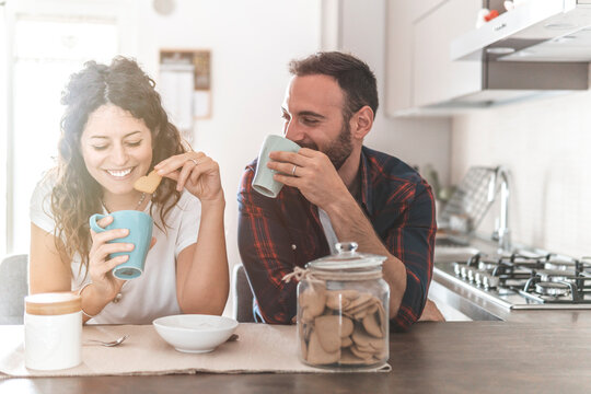 Engaged couple has breakfast together in their new home - young couple smiling while drinking and eating in the kitchen - warm filter on background