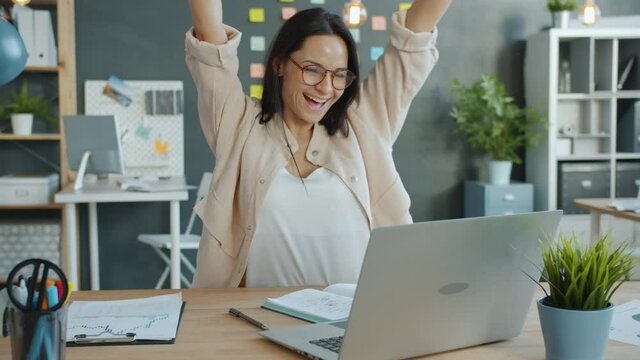 Joyful business lady is enjoying successful work with laptop having fun getting good news in workplace raising arms and smiling. People and success concept.