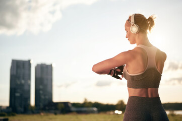 Back view of strong muscular woman in sportswear and headphones is cheking smartwatch and using...