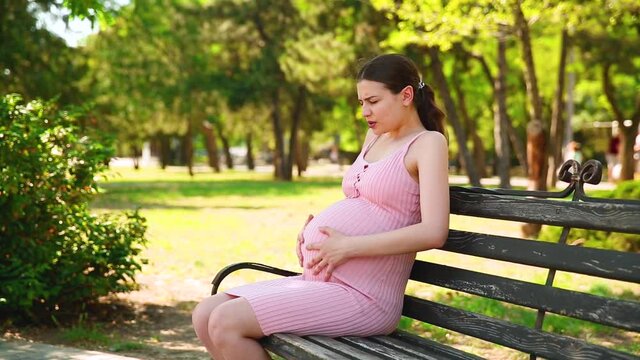 A pregnant girl is sitting on a park bench, and her stomach begins to hurt and she begins to breathe correctly. pregnant woman stroking belly