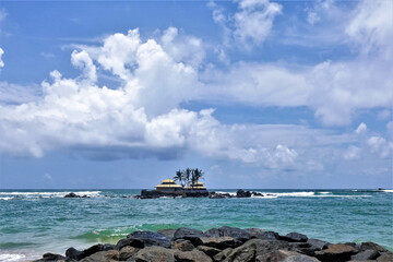 Naklejka premium An amazing Buddhist temple in the Indian Ocean. On a tiny rocky island near the shore there is a small temple. Aquamarine waves beat against the rocks. Blue sky with picturesque clouds. Sri Lanka. 