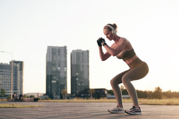 Strong athletic woman doing squats outdoor and listening to music in headphones, backlit sunset,...