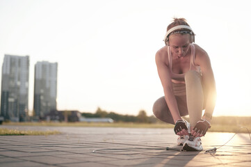 Strong athletic girl in sportswear and headphones tying shoelaces on sneakers on the street in the...