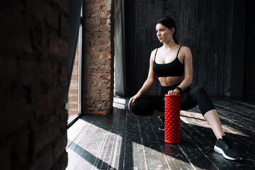 Sports shooting of a girl who sits on the floor in a loft and holds a massage red cylinder in front of her to study fascia