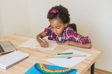 Afro child draws at a desk. Focused on work. 9 years old pre-teen. Nearby is laptop, notebooks, pencils, ruler, handmade rainbow. Distance learning, online education, homework, back to school concept.