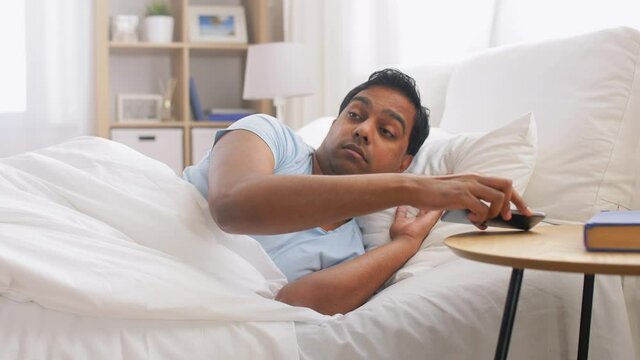 people, bedtime and rest concept - sleepy indian man in bed suddenly awaking and looking at smartphone lying on table at home