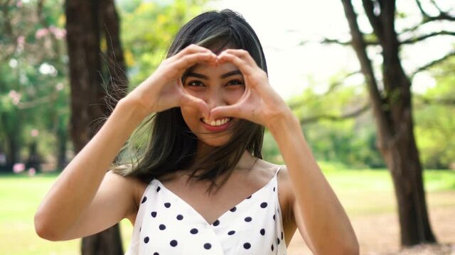 Young brunette woman with smile face making heart shape hands gesture in park. Beautiful Asian girl with toothy smile outdoors.
