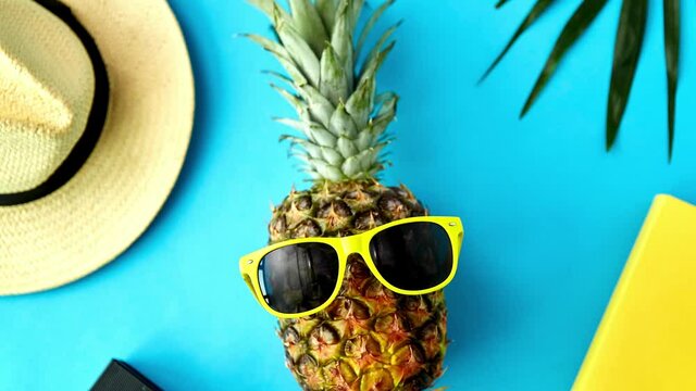 summer holidays, vacation and travel concept - close up of pineapple in yellow sunglasses, hat, camera and palm leaf on blue background