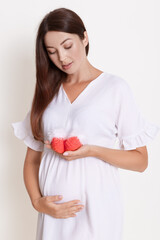 Beautiful pregnant woman with red baby shoes isolated on white background, lady expecting baby looking at small booties in her hand and touching her belly.