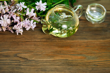 Herbal tea in a glass cup and teapot with bouquet of saponaria flowers on a wooden background. Copy space.