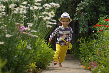 Summer portrait of little smiling boy in straw hat and bright clothes running fast along pathway among huge flowers