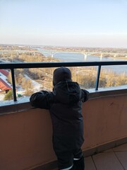 A small boy stands on the balcony in the entrance and looks at the snow covered city on the river Bank