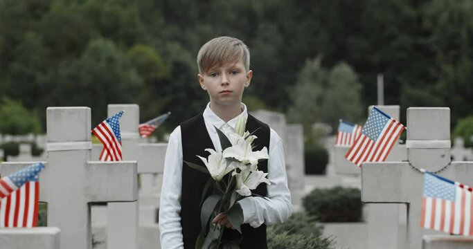 Portrait of teenger child looking to camera while standing in row of stone crosses with american flags on them. Young boy holding white lily flower at cemetery. Concept of memorial day