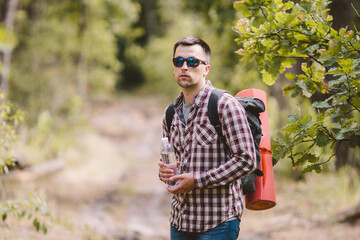 Naklejka na ściany i meble Hiker Drinking Water In Forest. Tired man drinking water from bottle in woodland. Handsome Traveler With Backpack And Flask In Forest. vacation, outdoor lifestyle freedom concept. Adventures hiking