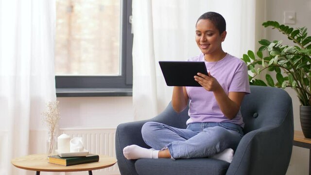 people, technology and leisure concept - happy young african american woman with tablet pc computer sitting in chair at home