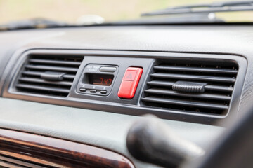 Closeup of a red emergency light button with a white triangle between the air conditioning grilles on the dashboard in the interior of a car.