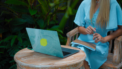 A girl in a blue dress remotely online working behind a laptop in the backyard with green plants on the background