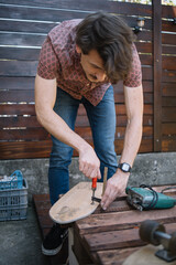 Man with screwdriver fitting up skateboard with tires. Man's hands making skateboard and using screwdriver on table outdoor.