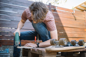 Handyman using power saw on skateboard deck . Human using power hand saw while making skateboard with wooden wall in background.