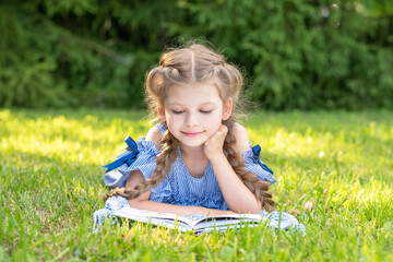A little girl reads a book lying on a green lawn in the Park. Outdoor reading.