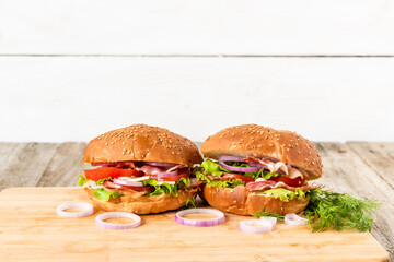 Two sandwiches with lettuce, bacon, onion rings, tomatoes, egg, cheese and a sesame bun on a wooden background.