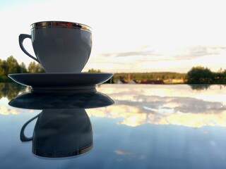 White tea cup on a glass table. The mirror surface reflects morning light and a cup and saucer.