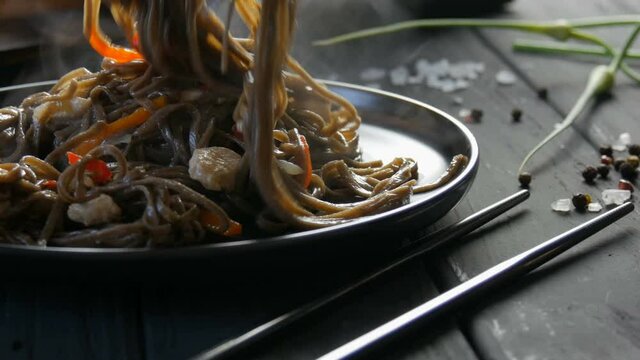 Put in beautiful stylish black plate Freshly boiled steam asian buckwheat noodles soba with vegetables onion pods, carrots, paprika and pieces of chicken near Chinese chopsticks.